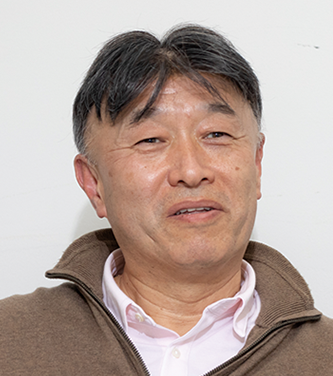 Portrait of Mr. Matsumura, an adult man in a collared shirt and brown sweater, shown against a plain background.