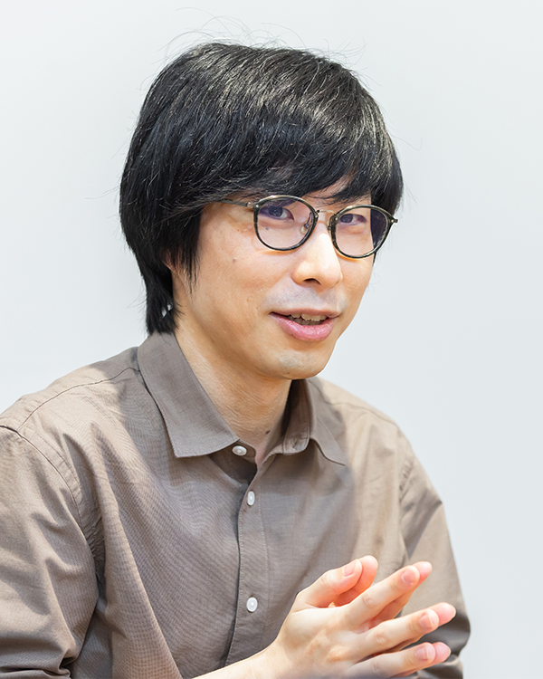 A person with black hair, glasses, and a brown shirt, sitting and looking towards the camera with a neutral expression.