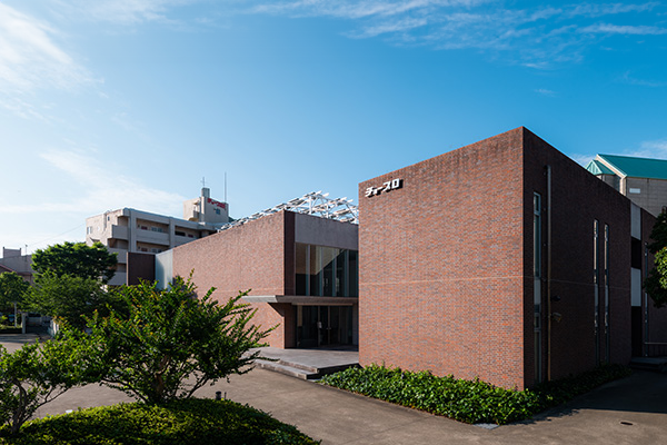 The exterior view of a modern brick building with 'チョープロ' signage, surrounded by greenery, under a blue sky during daytime.
