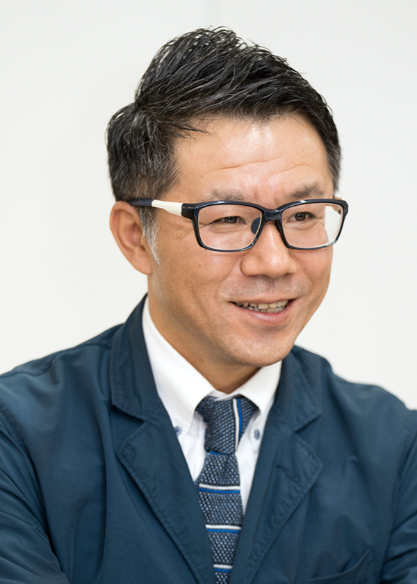 A person wearing a navy blazer, white shirt, and patterned tie with neatly styled dark hair.