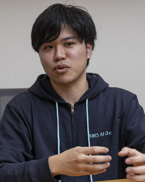 A person wearing a dark hoodie that reads 'AWS AI Day' is seated at a table, gesturing with their hands while speaking. This image is part of the Saison Technology case study interview.
