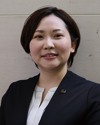 A professional headshot portrait of a person in business attire standing against a light-colored wall.