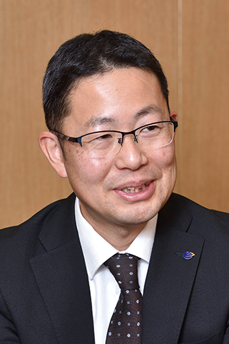 Portrait of a Meitetsu executive wearing a black suit, white shirt, patterned tie, and glasses, posing in front of a neutral background.