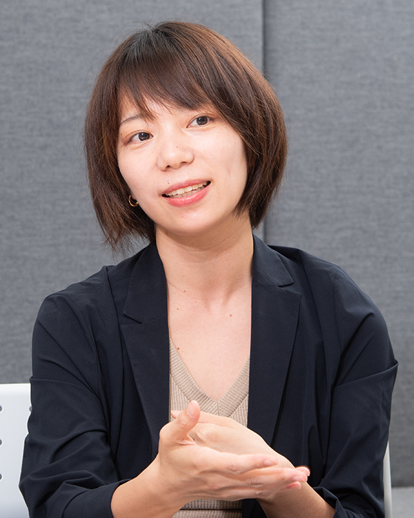 A person sitting indoors during an interview, wearing a dark blazer and light top, with a neutral background. This image is used for the ExaWizards case study featuring an interview with Ooaza Saori.