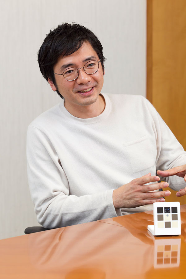 A man in glasses and a white sweater sits at a conference table, speaking and gesturing during an interview, with a color calibration card on the table in front of him.