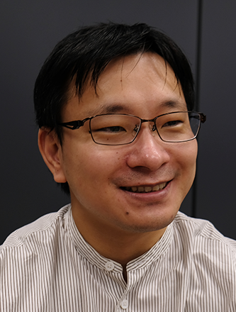 A smiling man wearing glasses and a light-striped shirt, photographed against a dark background.