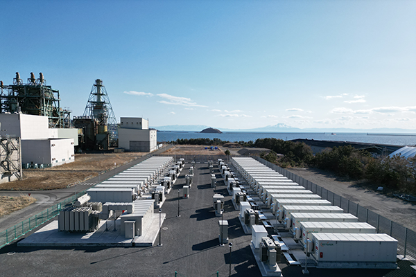A wide view of the ENEOS industrial energy storage facility in Japan, featuring multiple large battery storage units arranged in rows within a fenced area, with industrial infrastructure and the sea in the background.