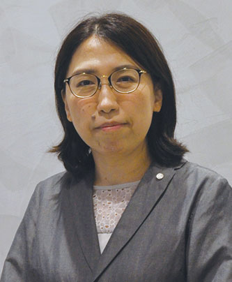 A person wearing a gray blazer over a patterned blouse, standing against a neutral background.