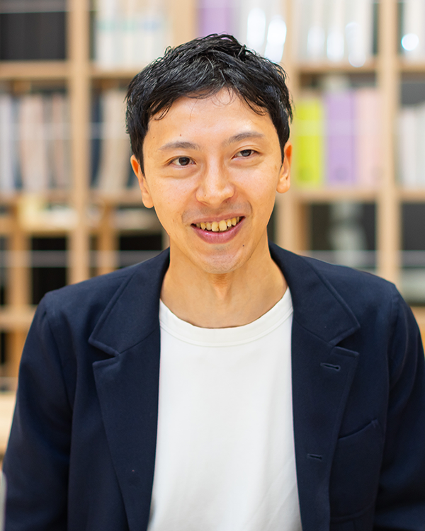 A portrait of a smiling man in a dark blazer and white shirt, standing in front of a blurred bookshelf background. Used for the ENEOS case study.