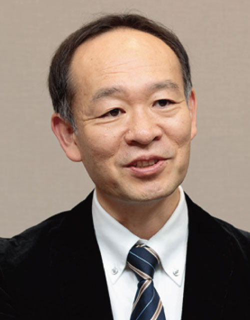 A person wearing a black suit jacket, white collared shirt, and striped tie against a beige background.