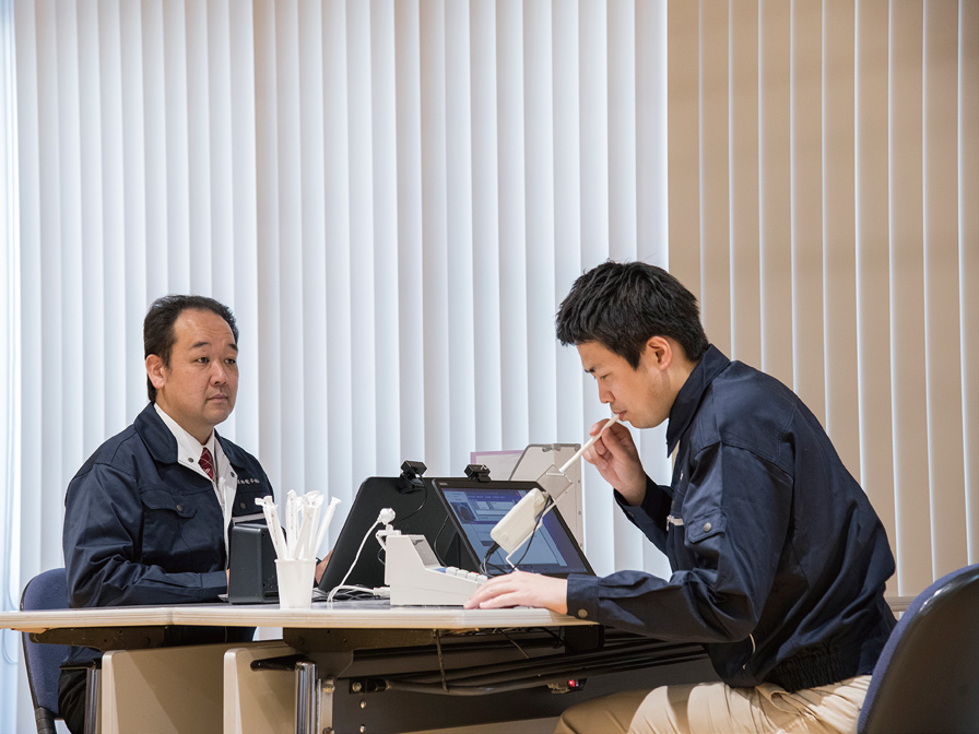Two individuals seated at a desk with electronic equipment and white blinds in the background.