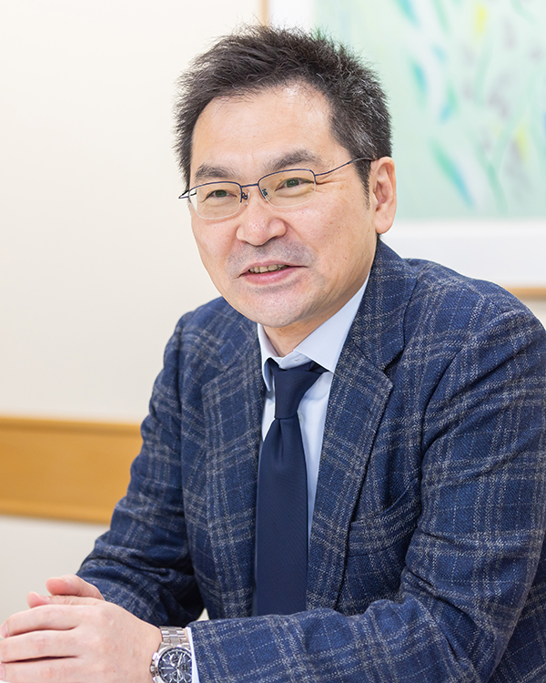 A man in a blue checkered suit and glasses is sitting at a table, speaking in an interview setting. The image appears to be from an interview with NTT East Yamaguchi, with a light background and framed artwork visible behind him.