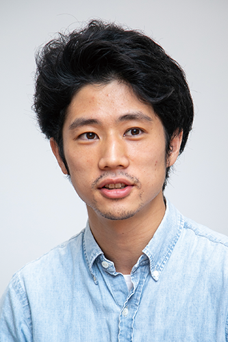 Portrait photograph of a man with dark hair wearing a light blue button-up shirt, facing forward in a neutral indoor setting.