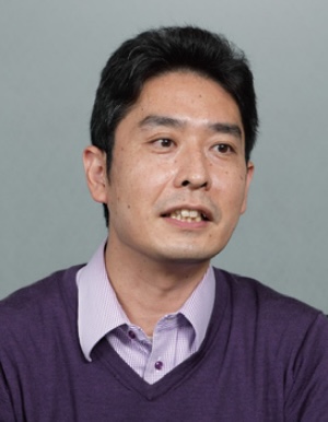 A person wearing a purple sweater over a collared shirt against a gray background.