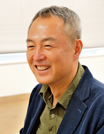 A smiling man wearing a green shirt and blue blazer sitting indoors.