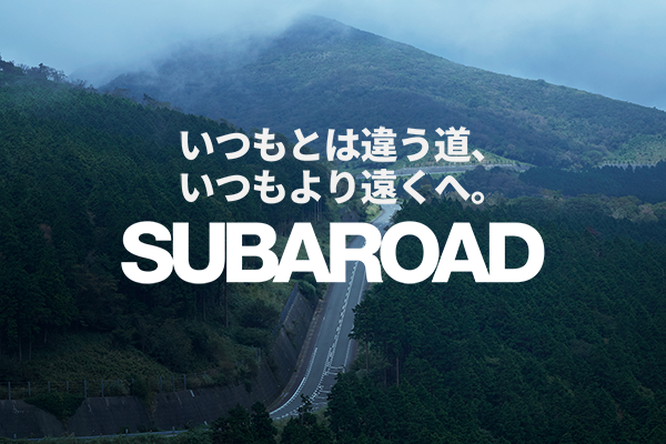 A scenic mountain road passing through a lush green forest with the Subaru campaign slogan in Japanese, reading 'いつもとは違う道、いつもより遠くへ。SUBAROAD', which translates to 'A different road than usual, farther than ever. SUBAROAD.'