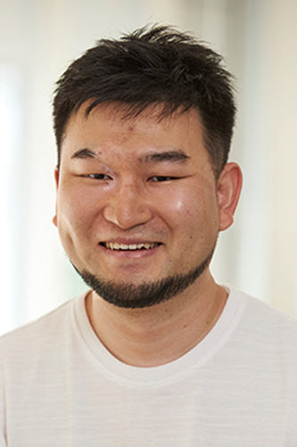 Portrait of a smiling man with short hair and a trimmed beard, wearing a white shirt, photographed against a softly blurred indoor background.