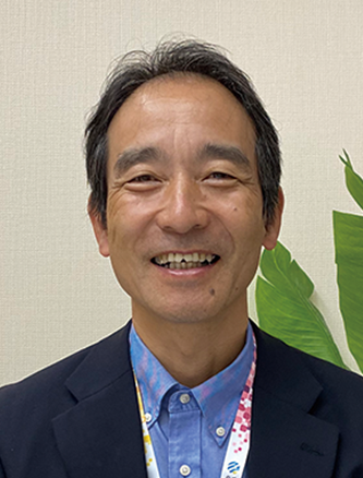 Portrait of a smiling man in business attire, photographed indoors with a plant in the background.