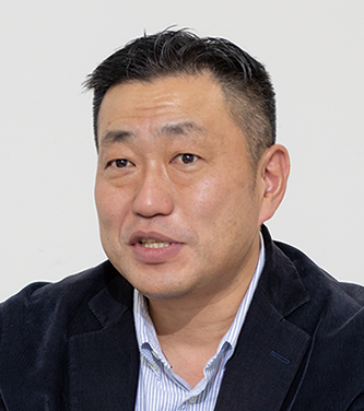 Portrait of a man in a dark blazer and striped shirt, facing the camera against a plain background.