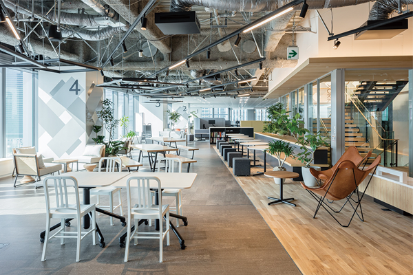A modern open-plan office interior with work tables, lounge chairs, plants, and exposed ceiling, featured in the Itoki AWS case study.
