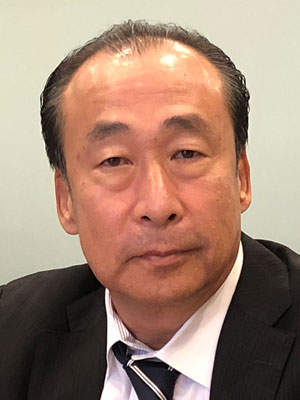 A person wearing a suit, white shirt, and striped tie against a plain background.