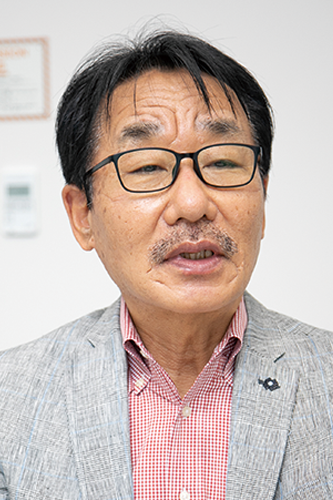 Portrait photo of a man wearing glasses and a gray suit jacket over a red checkered shirt, photographed against a light background.