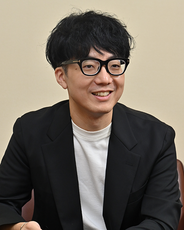 Portrait of a man with black hair and glasses, wearing a black jacket and white shirt, smiling and seated. Image used for Takihyo case study on the AWS Japan website.