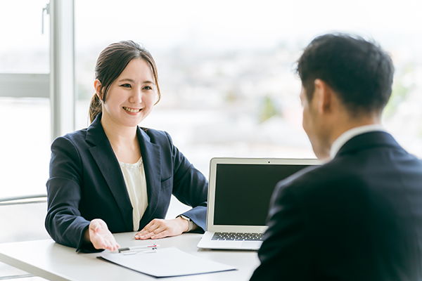 A professional business meeting with two people in formal attire, one woman smiling and gesturing towards a clipboard, and a laptop on the table in a bright office setting.