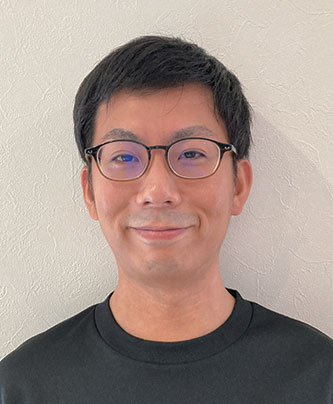 A portrait of a man wearing glasses and a black shirt, standing against a light-colored textured wall.
