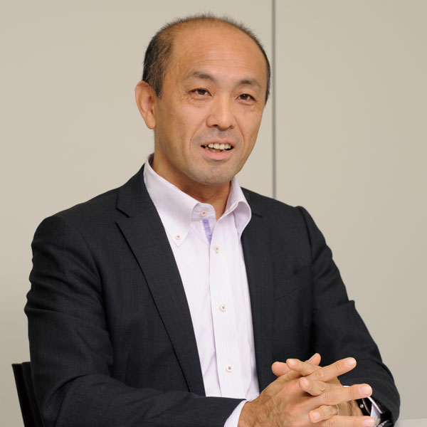 A man in a business suit, seated and speaking during an interview, set against a neutral background. Ideal for use in business, interview, or case study contexts.