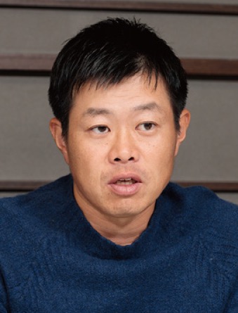 A man wearing a blue sweater is talking indoors, with a blurred background featuring horizontal lines.