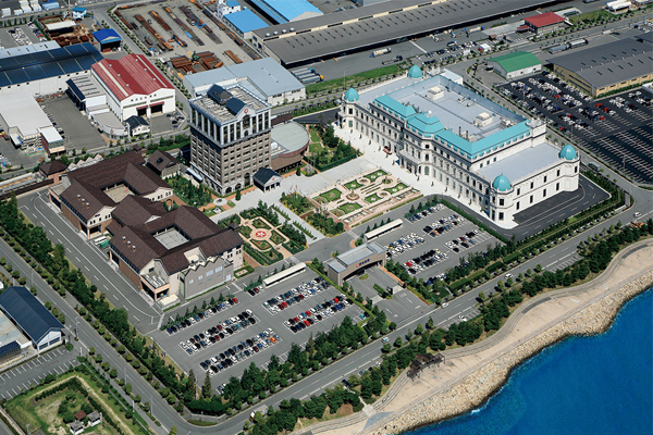 Aerial view of the Nihon Shokken facility, showing multiple buildings, parking lots, landscaped gardens, and its proximity to the waterfront.