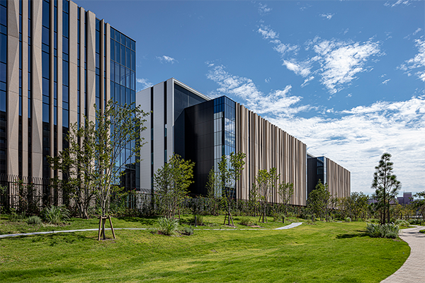 Modern Chugai Pharmaceutical headquarters building with glass and vertical panel architecture, landscaped with green lawn and young trees under a blue sky.