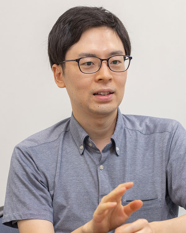 A portrait of a man with short dark hair and glasses, wearing a gray collared shirt, speaking or gesturing with his hands. Neutral background.