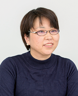 Portrait of a woman with short hair, wearing glasses and a navy sweater, seated and smiling against a plain background.