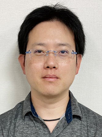 Professional portrait of an individual with short black hair, wearing glasses and a grey collared shirt, photographed against a light background.