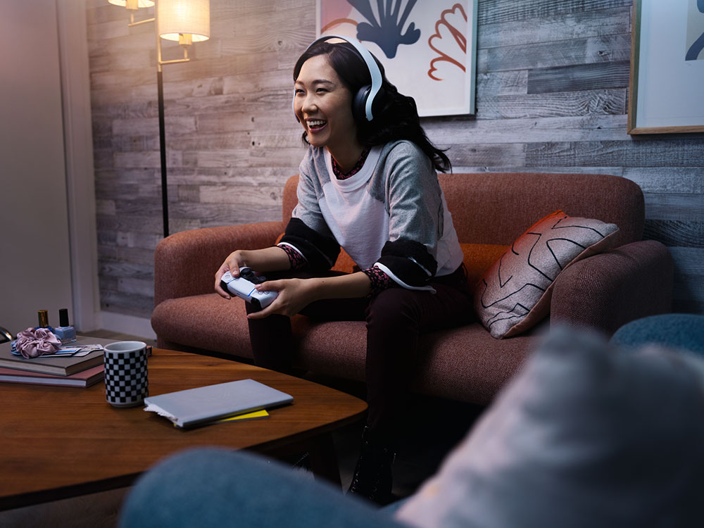A woman wearing headphones is sitting on a couch while playing a video game with a controller, with a cozy living room setting in the background.