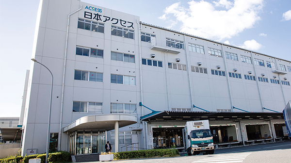 The exterior view of a large, modern white building labeled 日本アクセス (Japan Access), with several floors and glass windows. There is a truck parked in front and a blue sky with clouds in the background.