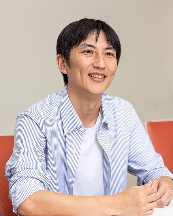 A person wearing a blue and white striped shirt over a white t-shirt, seated at a table with an orange chair in the background.