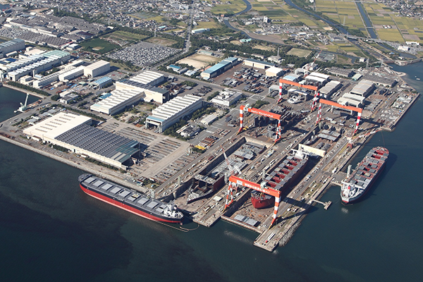 Aerial view of a large shipyard with multiple docks, cranes, and ships under construction or repair, surrounded by industrial buildings and a nearby town.
