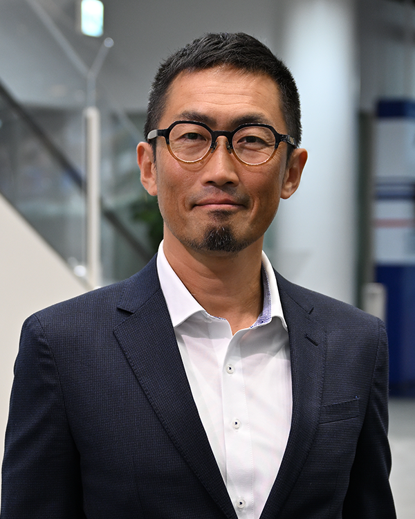 A person wearing a dark suit and white shirt standing indoors with a modern office background.