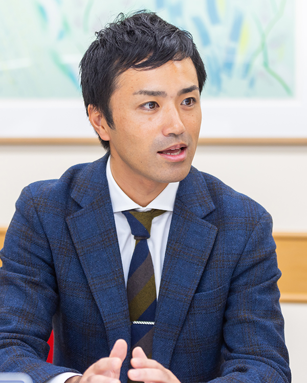A person wearing a blue plaid suit, white shirt, and striped tie, seated in front of a blurred abstract background.