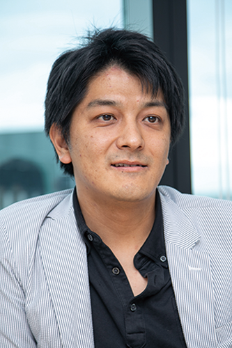 Portrait of an individual in a business setting from the Softbrain ODA interview, wearing a striped jacket and black shirt, seated indoors with background window view.
