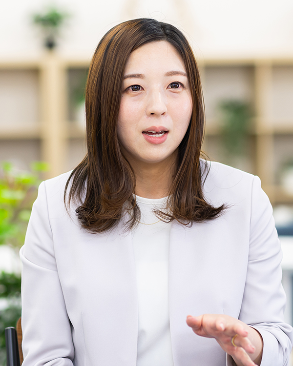 A woman in a white blazer is speaking during an interview, set in a modern office environment with shelves and plants in the background. 
