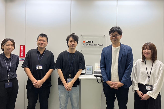 A group of five people standing in front of a wall with an "Orico" sign and office equipment visible.