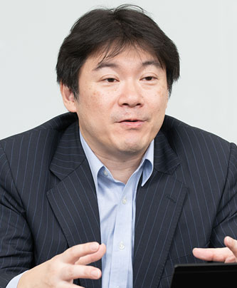 A portrait of a man in a business suit and light blue shirt, speaking and gesturing with his hands. The background is plain and light colored.