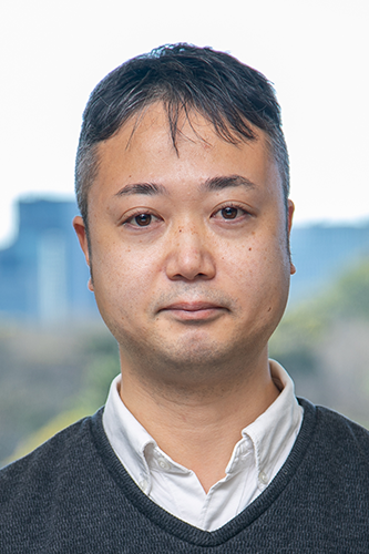 A portrait photograph of a man wearing a dark sweater over a white collared shirt, with a blurred outdoor background.