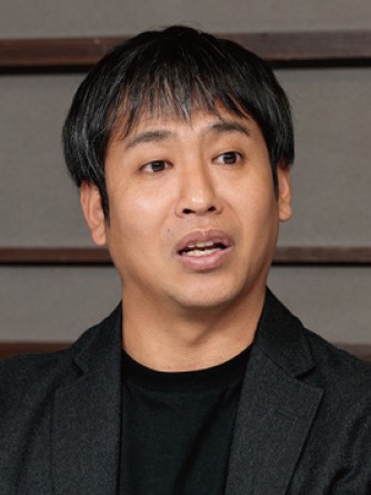 An Asian man with black hair, wearing a black blazer and black shirt, talking while facing slightly to the side.
