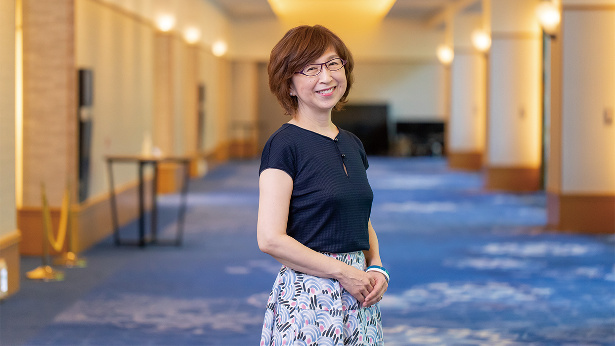 A woman with short hair and glasses is smiling while standing in a hallway. She is wearing a navy top and a patterned skirt, and the background shows a carpeted hallway with warm lighting.