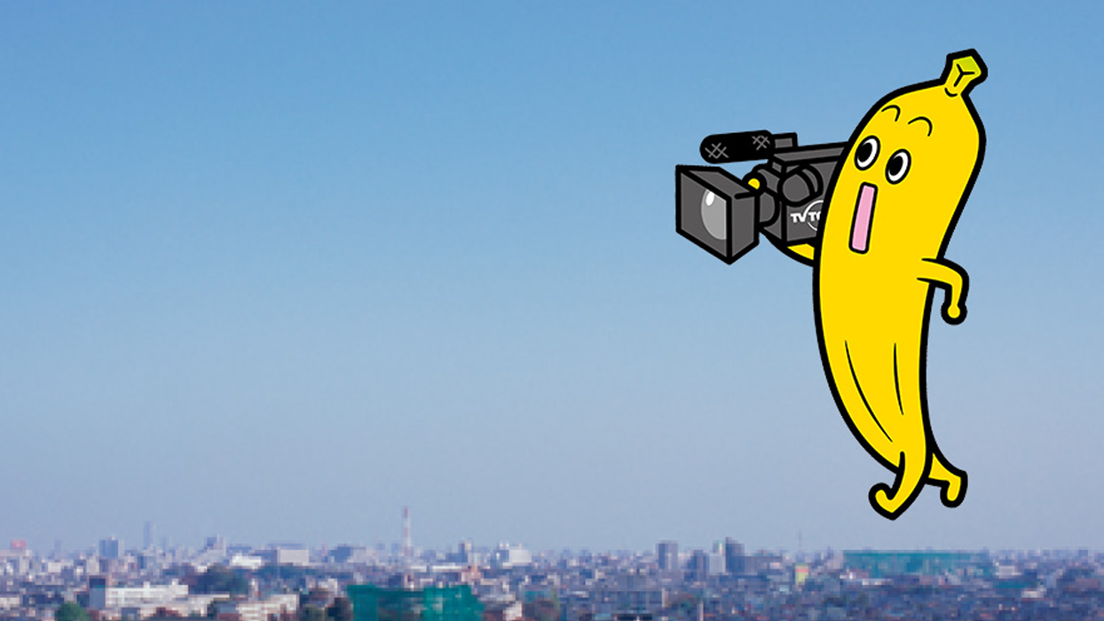 An illustrated yellow character resembling a banana holding a video camera, with a cityscape and clear blue sky in the background. Used for TV Tokyo Python tutorial content.